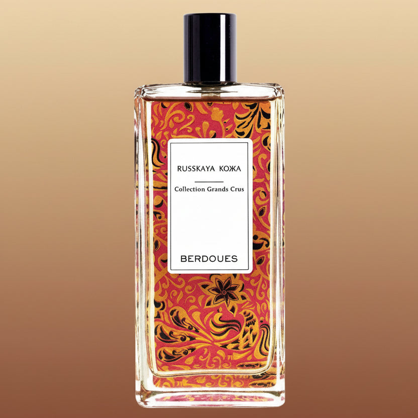 Berdoues Russkaya Кожа tall rectangular perfume bottle with a red, orange and black design on it, a white label with black lettering, a black cylindrical cap on a warm gradient background.